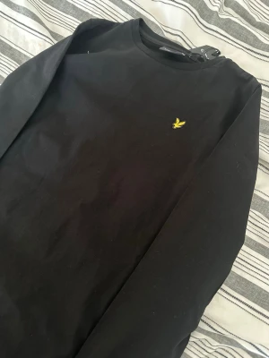 Svart långärmad tröja Lyle & Scott - Säljer en svart långärmad tröja från Lyle & Scott med den klassiska gula loggan broderad på bröstet. Tröjan har rund halsringning och är tillverkad i mjukt bomullsmaterial. Enkel och stilren design som passar till det mesta.
