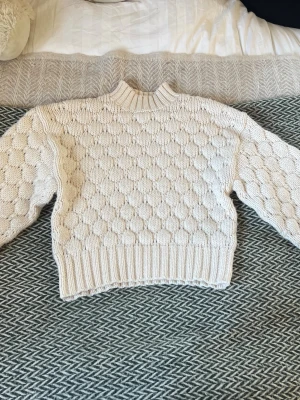 Vit chunky stickad tröja - Säljer en vit chunky stickad tröja med strukturmönster och ribbade muddar vid ärmslut och nederkant. Tröjan har en högre halsringning och är gjord i ett tjockare garn, perfekt för kyliga dagar.