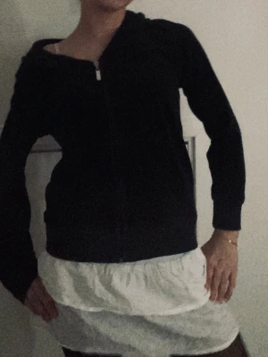 Svart hoodie med dragkedja - En svart hoodie med dragkedja framtill och huva. Klassisk modell med långa ärmar och ribbade muddar vid ärmslut och nederkant. Perfekt för en avslappnad och stilren look.