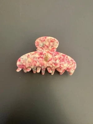 Mönstrad hårklämma i rosa och vitt - Säljer en snygg hårklämma med ett unikt mönster i rosa, vitt och lite rött. Klämman har en klassisk form och är tillverkad i plast. På ena sidan finns en liten guldfärgad detalj som ser ut som en ananas. Perfekt för att snabbt fixa frisyren.