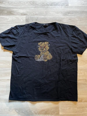 Svart t-shirt med nalletryck - Svart t-shirt med ett stort tryck av en nallebjörn i guld och silver på bröstet. T-shirten har rund halsringning och korta ärmar. Perfekt för dig som vill ha en cool och lekfull stil.