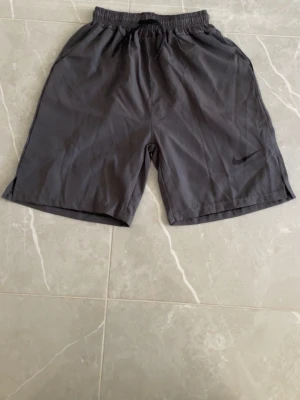 Grå träningsshorts från Nike - Säljer ett par grå träningsshorts från Nike med elastisk midja och snörning. Shortsen har en enkel design med Nike-logga på benet och är tillverkade i ett lätt och bekvämt material som passar perfekt för sport och aktivitet.