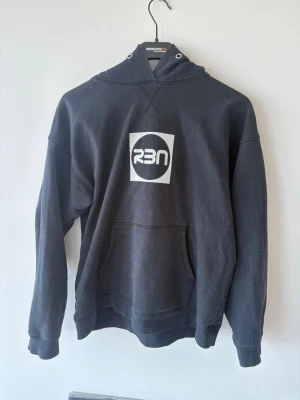 Svart hoodie med RBN-tryck - Säljer en svart hoodie från RBN med stor vit logga framtill och coolt grafiskt tryck på ryggen. Hoodien har huva med dragsko och en klassisk magficka. Tillverkad i mjukt bomullsmaterial som är skönt att ha på sig. Perfekt för dig som gillar streetwear och enkel stil.