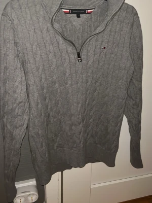 Grå kabelstickad tröja Tommy Hilfiger - Snygg grå kabelstickad tröja från Tommy Hilfiger med halv dragkedja och ribbad krage. Liten logga på bröstet och klassiska Hilfiger-detaljer vid dragkedjan. Perfekt till jeans eller chinos för en clean look.