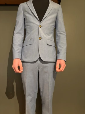 Ljusblå kostym från dressman - Snygg ljusblå kostym med klassisk kavaj och matchande byxor. Kavajen har två guldiga knappar, smala slag och fickor med lock. Kostymen har en modern, slim passform och är tillverkad i ett lätt material som passar perfekt till vår och sommar.