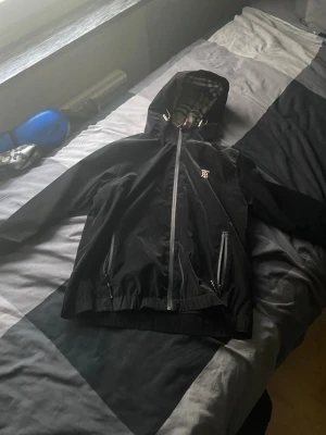 Svart vindjacka från Burberry - Snygg svart vindjacka från Burberry med klassiskt rutigt foder i huvan. Jackan har dragkedja framtill, två fickor med dragkedja och en liten Burberry-logga på bröstet. Perfekt för dig som vill ha en stilren och ikonisk look.