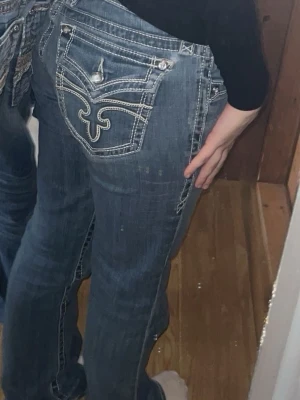 Rock Revival blå jeans med brodyr - Säljer ett par blå jeans från Rock Revival med snygga kontrastsömmar och broderad bakficka. Jeansen har bootcut, låg midja och detaljerade knappar. Tyvärr har fickorna gått sönder och har sytt dem men dem har gått sönder lite igen, men kan man sy tror jag absolut att dem går att rädda, skriv gärna innan köp.