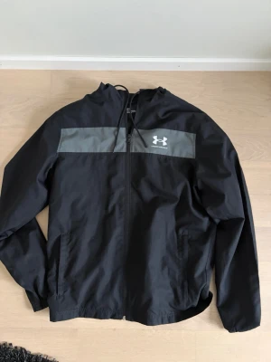 Svart vindjacka från Under Armour. (kolla beskrivning) - Snygg svart vindjacka från Under Armour med grått bröstparti och logga. Jackan har huva med dragsko, hel dragkedja framtill och två sidofickor. Tillverkad i lätt och tåligt material, perfekt för blåsiga dagar. Köp gärna med instabox. Skickar då samma dag om du har köpt den innan 16:00.