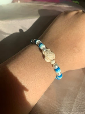 Pärlarmband med sköldpadda - Säljer ett pärlarmband med vita, blå och silverfärgade pärlor samt en söt sköldpaddsdetalj i mitten. Armbandet har en ungdomlig och strandig vibe och är elastiskt för enkel påtagning. Perfekt accessoar för dig som gillar unika detaljer.
