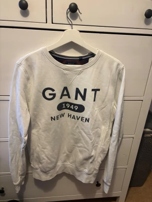 Vit sweatshirt från Gant - Vit sweatshirt från Gant med trycket 'GANT 1949 NEW HAVEN' i svart på bröstet. Tröjan har rund halsringning och långa ärmar. Perfekt för en avslappnad och stilren look.
