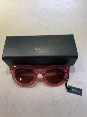 Rosa solglasögon från Mulberry - Säljer ett par snygga solglasögon från Mulberry i transparent rosa acetat med ovala bågar och tonade linser. Diskret Mulberry-logga på skalmen och medföljande svart fodral. Perfekt accessoar för dig som vill sticka ut med stil.
