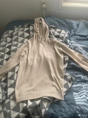 Beige hoodie med dragsko - Snygg beige hoodie med huva och dragsko. Tillverkad i ett mjukt, stickat material som känns skönt mot huden. Långa ärmar och ribbade muddar vid ärmslut och nederkant ger en clean look. Perfekt för dig som gillar stilrena basics. Stl 176