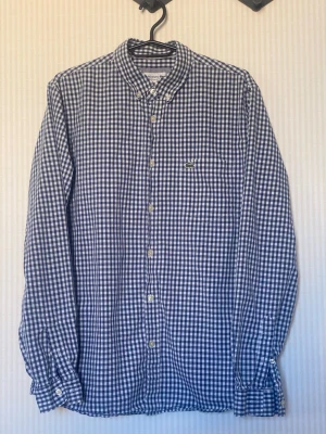 Blårutig skjorta från Lacoste - Klassisk blårutig skjorta från Lacoste med button-down krage och lång ärm. Skjortan har vita knappar framtill och en liten broderad krokodil på bröstet. Tillverkad i mjuk bomull som känns skön mot huden. Perfekt för en avslappnad men ändå stilren look.