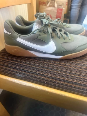 Nike gröna sneakers med vit swoosh - Snygga sneakers från Nike i olivgrönt och grått med vit swoosh på sidan och gummifärgad sula. Skorna har klassisk låg profil, snörning och är tillverkade i en mix av textil och syntetmaterial. Perfekta för en avslappnad streetstyle-look.