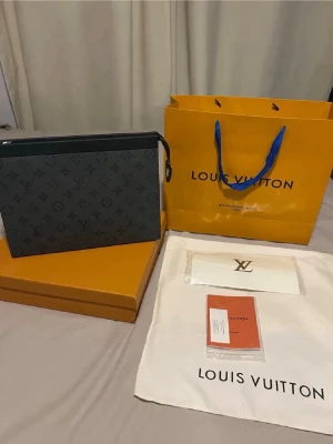 LOUlS VUlTTON POUCHETTE VOYAGE MM - Säljer en Voyage MM i det klassiska Monogram Eclipse (svart/grå). En otroligt mångsidig och snygg accessoar som fungerar perfekt som clutch eller rese-väska.  Modell: Pochette Voyage MM (M61692). Skick: Ny Mått: 27 x 21 x 3 cm. Detta medföljer (Fullset): Originalkartong & Dustbag & Originalkvitto.     Hör gärna av dig om du vill ha fler bilder eller har frågor! Frakt: Skickas spårbart och försäkrat