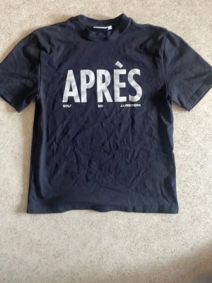 Svart t-shirt från J.Lindeberg - Snygg svart t-shirt från J.Lindeberg med stor vit text 'APRÈS' framtill. Klassisk rund halsringning och korta ärmar. Tillverkad i mjuk bomull som känns skön mot huden. Perfekt för en avslappnad och trendig look.
