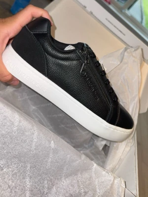 Svarta Calvin Klein sneakers med dragkedja - Snygga svarta sneakers från Calvin Klein Jeans med vit sula och dragkedja på sidan. Skorna är i slätt och strukturerat läder med svarta snören och diskret logga på sidan. Perfekta för en clean och modern stil.