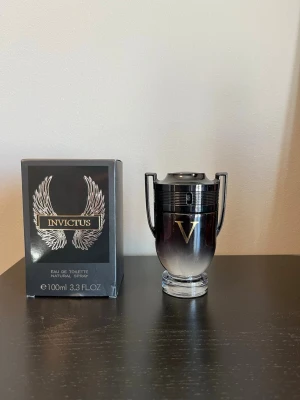 Paco Rabanne Invictus Eau de Toilette - Snygg parfymflaska från Paco Rabanne i form av en pokal, svart och silverfärgad med gulddetaljer och ett stort V på framsidan. Kommer i originalkartong med vingar och logga. Innehåller 100 ml Eau de Toilette spray.