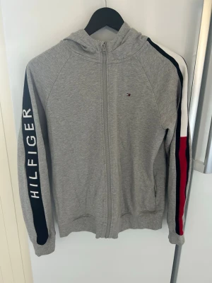 Grå hoodie från Tommy Hilfiger - Snygg grå hoodie med dragkedja från Tommy Hilfiger. Har huva, ribbade muddar och fickor framtill. Ärmarna har marinblå, vita och röda detaljer samt stort HILFIGER-tryck på ena ärmen. Liten broderad logga på bröstet. Perfekt för en chill och sportig look.