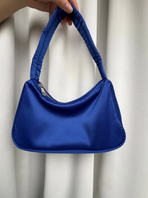 Blå axelväska i satin - Snygg blå handväska med axelrem i matchande tyg och mjuk, glansig satin-look. Väskan har en enkel, avlång form och stängs med dragkedja i silver. Perfekt för dig som vill ha en trendig accessoar som sticker ut.