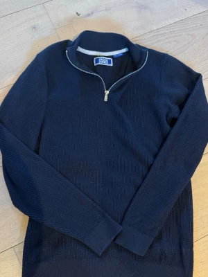 Mörkblå tröja med halv dragkedja - Mörkblå långärmad tröja från Jack & Jones med ribbad struktur och halv dragkedja vid halsen. Perfekt för lager på lager och enkel att matcha med jeans eller chinos. Klassisk och stilren design med mjukt material. Nyskick 