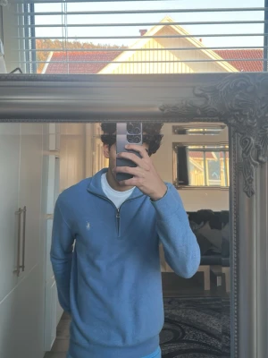 Blå halvzip-tröja från Polo Ralph Lauren - Snygg ljusblå stickad tröja från Polo Ralph Lauren med halv dragkedja och klassisk krage. Tröjan har lång ärm och den ikoniska broderade loggan på bröstet. Perfekt till jeans eller chinos för en clean och stilren look.