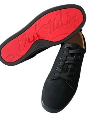Svarta Louboutin sneakers med röd sula - Snygga svarta sneakers från Christian Louboutin med ikonisk röd sula och diskret logga undertill. Skorna har en klassisk låg siluett, svarta skosnören och ovandel i mocka som ger en lyxig känsla. Perfekta för dig som vill sticka ut med stil.