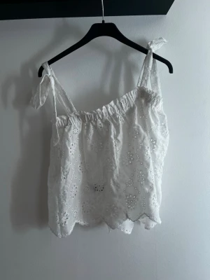 Vit broderad topp med knyt - Söt vit topp med broderade blommor och knytbara axelband. Toppen har en luftig passform och vågig kant nertill, perfekt för varma dagar. Romantisk och somrig vibe!