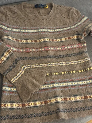 Brun mönstrad stickad tröja Polo Ralph Lauren - Säljer en brun stickad tröja från Polo Ralph Lauren med färgglada mönster i gult, rött, blått och vitt. Tröjan har rund halsringning, långa ärmar och detaljerade stickade mönster. Perfekt för dig som gillar retrovibbar och unika plagg.