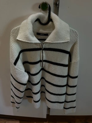 Vit stickad tröja med svarta ränder - Säljer en vit stickad tröja från Bik Bok med svarta horisontella ränder. Tröjan har en hög krage med dragkedja som kan bäras både uppvikt och nedvikt. Ribbstickad struktur och lång ärm för en avslappnad look.