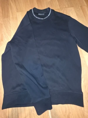 Svart sweatshirt från Emporio Armani - Stilren svart sweatshirt från Emporio Armani med diskret logga på bröstet och rund halsringning med vit textdetalj. Tröjan har långa ärmar och ribbade muddar vid ärmslut och nederkant. Perfekt för en clean och avslappnad look. Storlek S herr, använd ett fåtal gånger