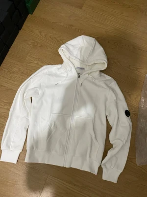Vit hoodie från C.P. Company - Säljer en stilren vit hoodie från C.P. Company med dragkedja framtill och två stora fickor. Klassisk huva och den ikoniska svarta linsen på ärmen. Tillverkad i mjukt bomullsmaterial som är skönt att bära. Perfekt för dig som gillar streetwear och enkel design.