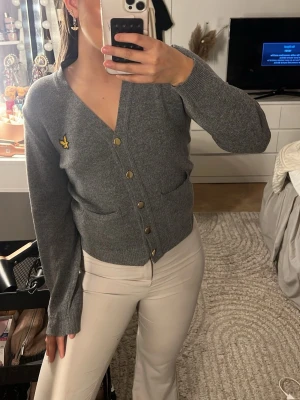 Grå kofta från Lyle & Scott - Snygg grå kofta från Lyle & Scott med gul logga på bröstet. Koftan har v-ringning, knappar framtill och två fickor. Tillverkad i mjukt stickat material och har långa ärmar. Köp på Sacon hand