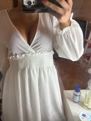 Vit omlottklänning med lång ärm - Säljer en vit kortklänning med omlott upptill och v-ringning. Klänningen har långa, lösa ärmar med resår vid ärmslut och smock i midjan för en snygg passform. Materialet känns lätt och luftigt, perfekt för varma dagar.