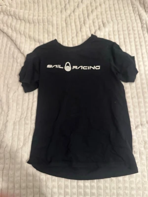 Svart Sail Racing t-shirt - Svart t-shirt från Sail Racing med vit logga tryckt över bröstet. Klassisk rund halsringning och korta ärmar. Skön och stilren modell som passar perfekt till jeans eller shorts. Materialet känns mjukt och bekvämt mot huden.det står 170 men det är för 150/155