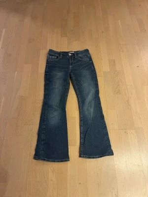 Blå bootcut jeans med detaljer - Snygga blå bootcut jeans med slitningar och dekorativa bakfickor med knappar. Jeansen har klassisk femficksmodell och kontrastsömmar. Perfekta för dig som gillar en avslappnad men trendig stil.
