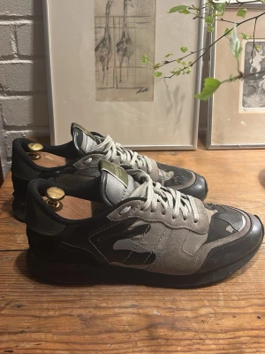 Valentino Rockrunners  - Snygga sneakers från Valentino med grå, svarta och olivgröna camo-detaljer. Ovandelen är i mocka och textil, med vita skosnören och robust gummisula. Märkeslogga på plösen och klassisk låg modell som passar till streetwear.