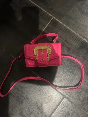 Rosa handväska med gulddetaljer - Snygg rosa handväska i konstläder med justerbar axelrem och dekorativt guldfärgat spänne framtill. Väskan har en strukturerad yta och ett handtag upptill för olika bärstilar. Perfekt accessoar för att lyfta din outfit.