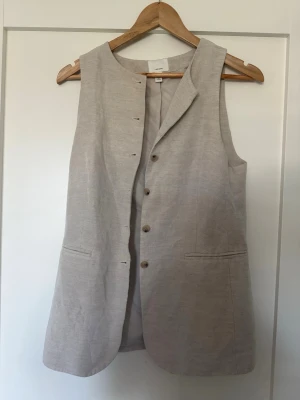 Beige lång linneväst från H&M - Stilren beige lång väst från H&M i linneblandning. Västen har knappar framtill, två fickor och en rak, avslappnad passform. Perfekt att slänga över en t-shirt eller skjorta för en clean look.  Vid kragen finns de en liten ljus gul fläck! Inget direkt synligt bara om man kommer för nära den! 