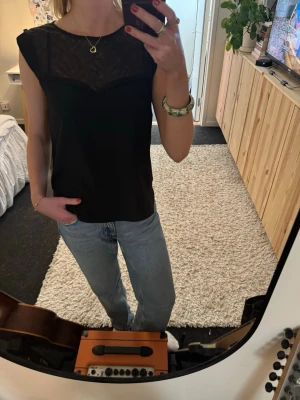 Svart ärmlös topp med spets - Snygg svart ärmlös topp med transparent spets upptill både fram och bak. Toppen har rund halsringning och knäppning i nacken. 