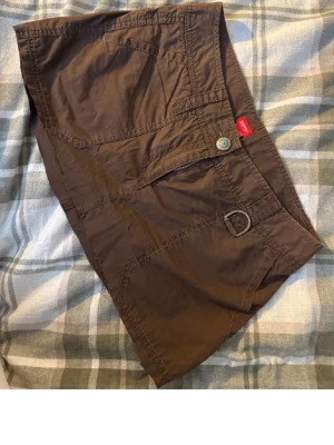 Brun utility skort från Esprit - Snygg brun skort från Esprit med flera fickor och dragkedjedetaljer. Skorten har en avslappnad utility-look med knappstängning och dekorativ D-ring i midjan. Tillverkad i bomull för en bekväm känsla och slitstark stil.