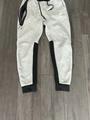 Nike grå och svarta mjukisbyxor - Säljer ett par grå mjukisbyxor från Nike med svarta detaljer längs benen och muddar vid bensluten. Byxorna har dragsko i midjan och fickor med dragkedja. Perfekta för chill eller träning, riktigt sköna och snygga.