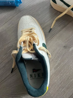 Nike vita och gröna sneakers - Snygga vita Nike sneakers med gröna detaljer och beige skosnören. Skorna har en klassisk låg siluett, perforerad tå och gummisula. Insidan är mörkgrön och det finns en gul detalj på hälen. Perfekta för dig som gillar retrostil och streetwear.