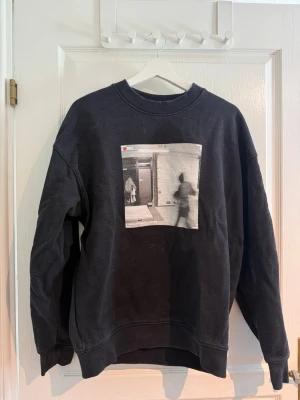 Svart sweatshirt med tryck - Cool svart sweatshirt med ett stort svartvitt tryck på bröstet som föreställer en suddig övervakningsbild. Tröjan har rund hals, ribbade muddar och avslappnad passform. Perfekt för dig som gillar streetwear och vill ha en unik look.