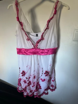 Y2K topp - Romantisk blus i vitt med rosa broderier, bred satinrosett i midjan och v-ringning. Ärmlös modell med volangkant nedtill och tunna axelband. Perfekt för dig som gillar feminina detaljer och unik stil. Storlek 12