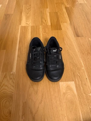 Svarta Puma sneakers i läder - Svarta sneakers från Puma med klassisk låg profil och snörning. Skorna är tillverkade i läder med diskreta loggor på sidan och tungan. Snygg och stilren design som passar till det mesta. Perfekta för dig som gillar en clean och sportig look.