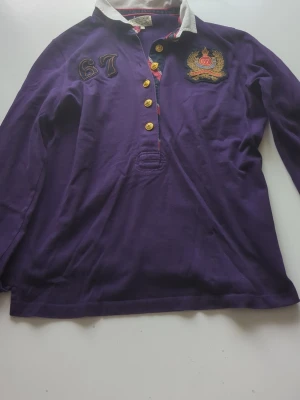 Lila gammal 67 Henley tröja - Säljer en lila henley-skjorta med lång ärm och guldfärgade knappar. Skjortan har vit krage, broderad detalj med siffran 67 och emblem på bröstet. Perfekt för dig som gillar sportig och preppy stil. Den är gammal men fortfarande i toppen skick