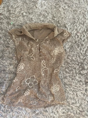 Brun / beige blommig blus  - Snygg brun blus med vita blommönster och prickig krage. Blusen har korta ärmar och knappar framtill. Perfekt för dig som gillar retrovibbar och vill ha något unikt i garderoben. Materialet känns lätt och luftigt.