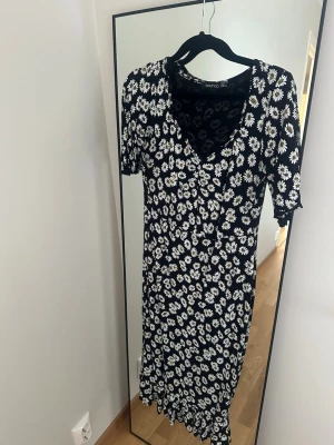 Blommig långklänning från Boohoo - Svart långklänning från Boohoo med vita blommor, v-ringad hals och korta ärmar. Klänningen har en figurnära passform och ett somrigt blommönster som ger en fräsch look. Perfekt för dig som gillar en enkel men snygg stil.
