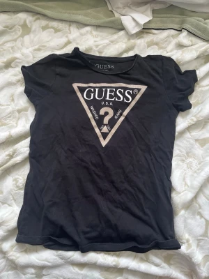 Svart t-shirt från Guess med logga - Snygg svart t-shirt från Guess med den klassiska triangelloggan i vitt och silver på bröstet. T-shirten har rund halsringning och korta ärmar. Perfekt för dig som gillar stilrena och ikoniska plagg.